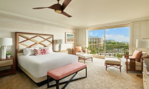 A resort guestroom featuring a large bed with a bench at the foot of it, two seating areas and an adjacent balcony